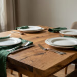 Table de salle à manger contemporaine inspirée des Arts and Crafts, dressée avec des assiettes blanches texturées, des couverts argentés martelés et du linge vert profond sous une lumière naturelle douce.